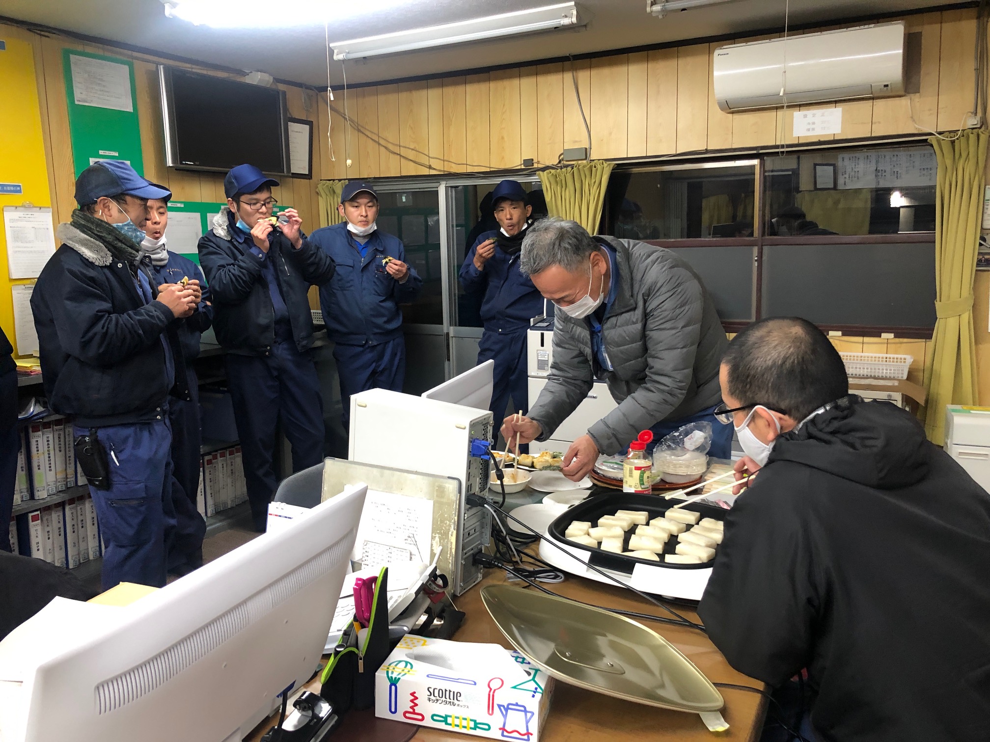 お餅を食べるサービスマンと、お餅を焼く社長と営業部長の写真