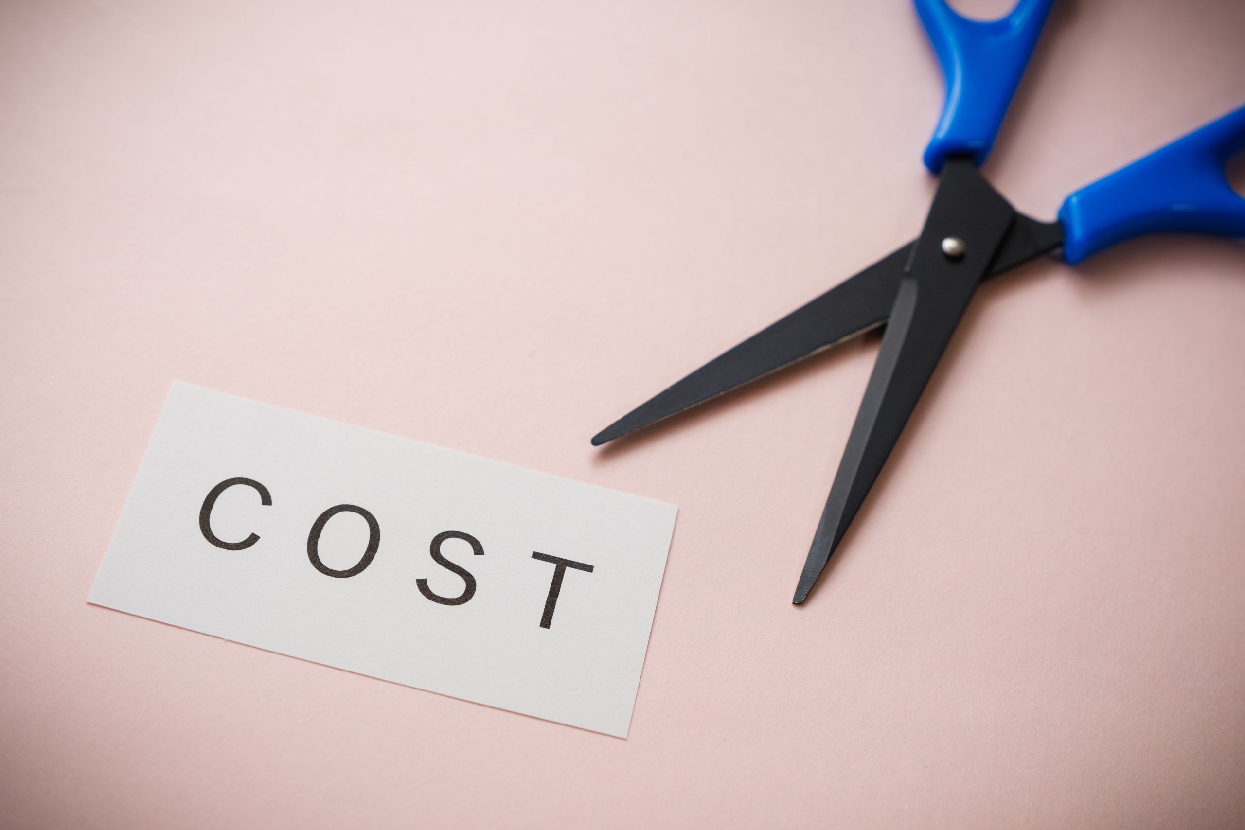 薄いピンク色の背景に「COST」と書かれた白い紙切れと、開いた状態の青いハサミが置かれている。ハサミの刃は「COST」の文字の近くにある。