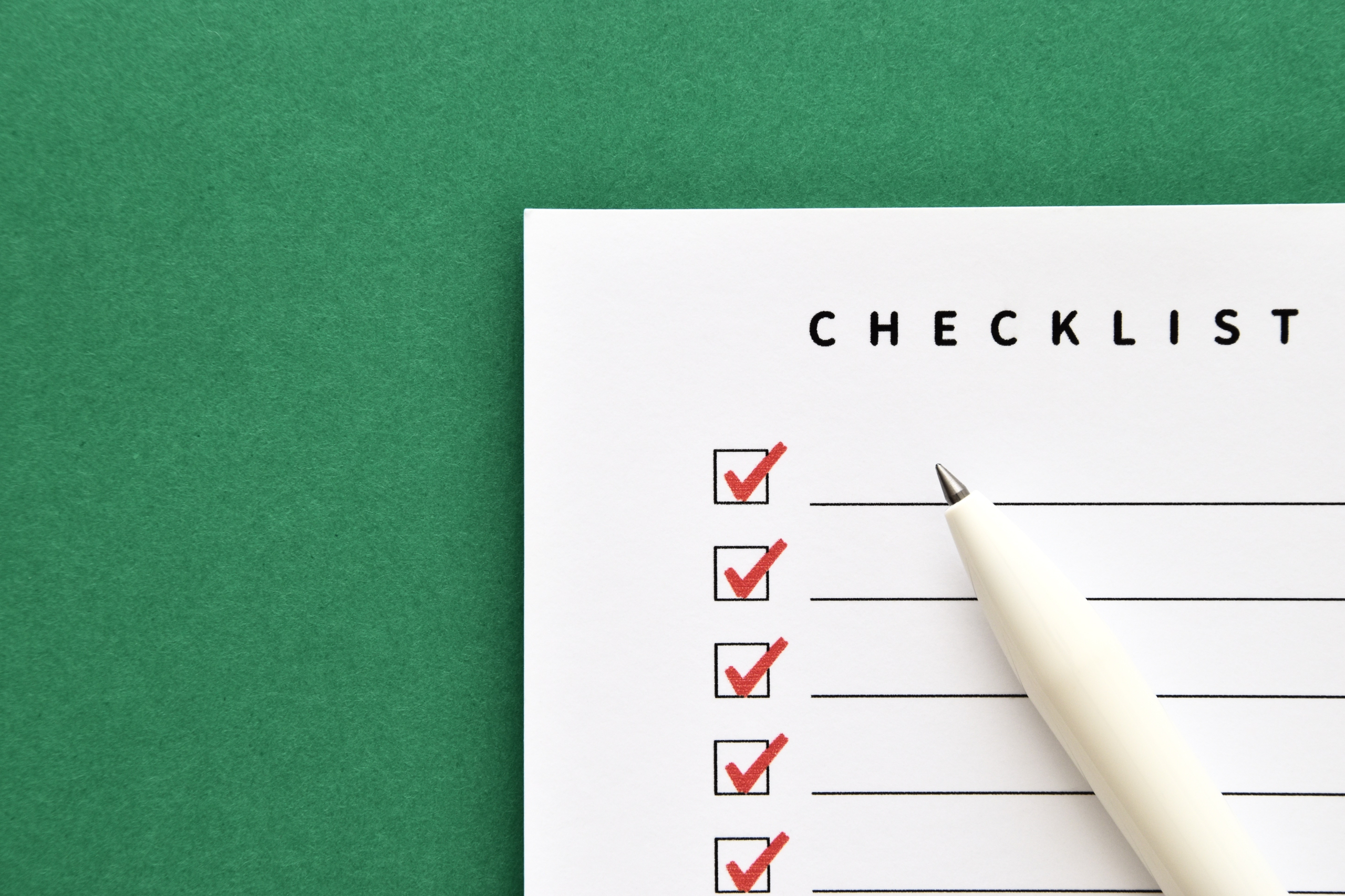 緑色の背景に、赤いチェックマークが入った「CHECKLIST」と書かれた白い紙と、その上に白いペンが置かれている。これは完了したタスクや計画を示すイメージ。