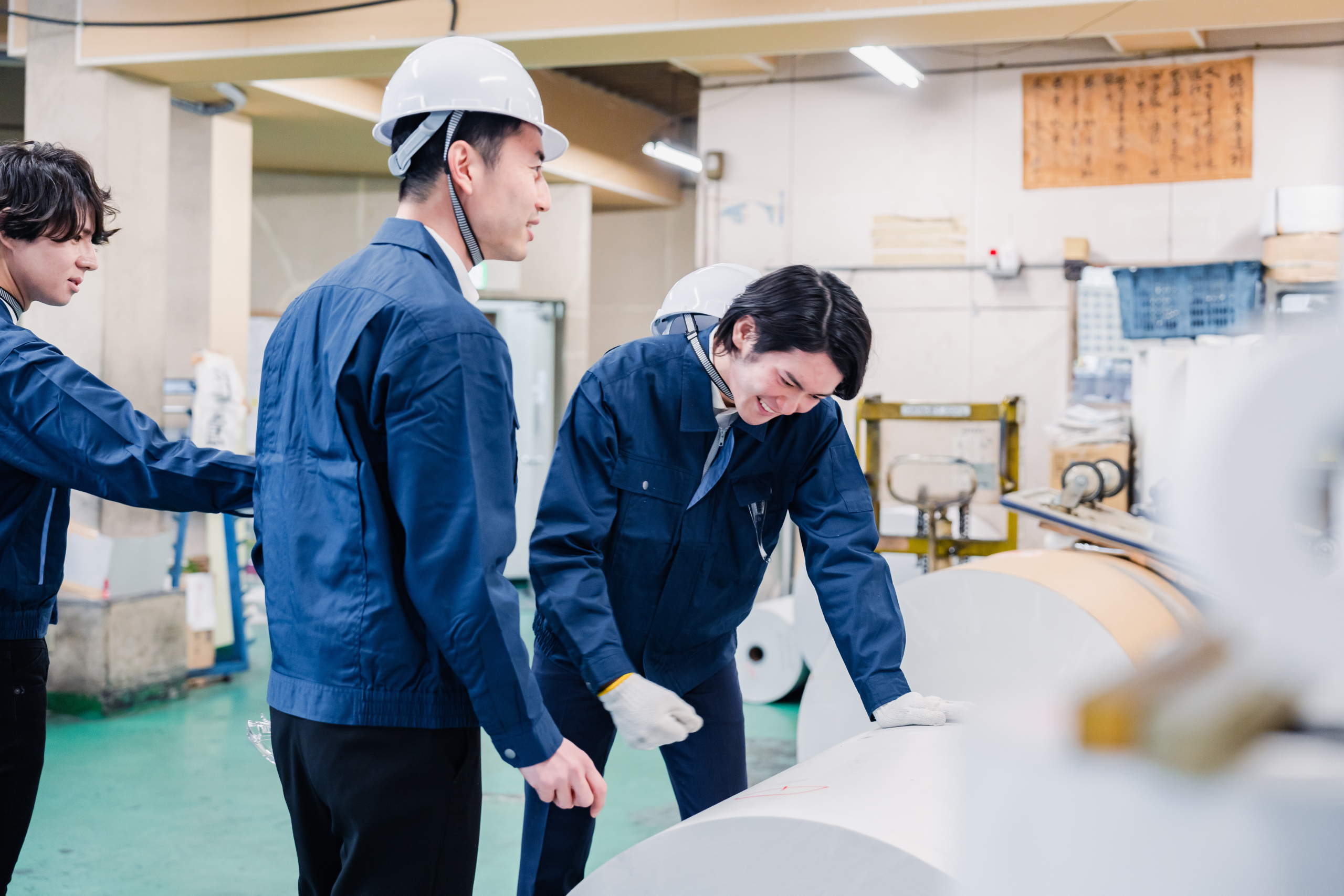 印刷工場で、青い作業服と白いヘルメットを着用した3人の男性作業員が、大きなロール状の素材を扱っている様子。彼らは協力して作業を進めており、真ん中の人物は屈んでロールに触れ、右の人物は笑顔で何かを話している。