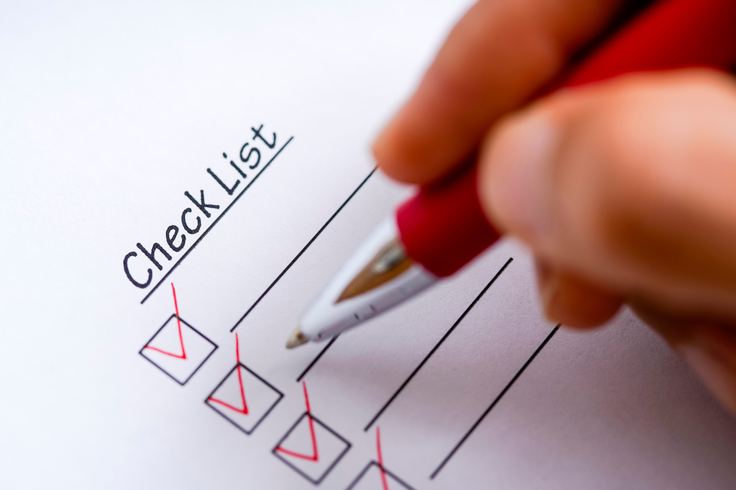 手が赤いペンで「Check List」と書かれた紙にチェックマークを入れているクローズアップ。いくつかの項目にはすでに赤いチェックが入っており、タスクの完了や確認作業の進行を示唆しています。