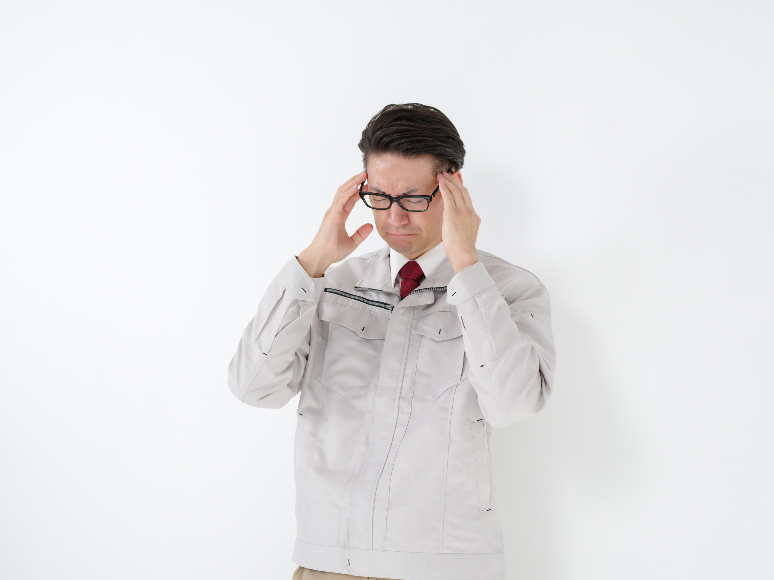 白い背景の前に立つ、白い作業服と赤いネクタイを着用した男性が、両手でこめかみを抑えて頭痛やストレスを抱えている様子。眼鏡をかけ、苦痛に顔をしかめています。